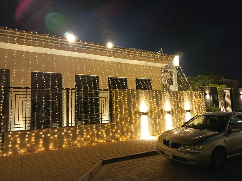 front view of a villa entrance decorated with curtain lights and bright wall lighting, with parked cars in front.