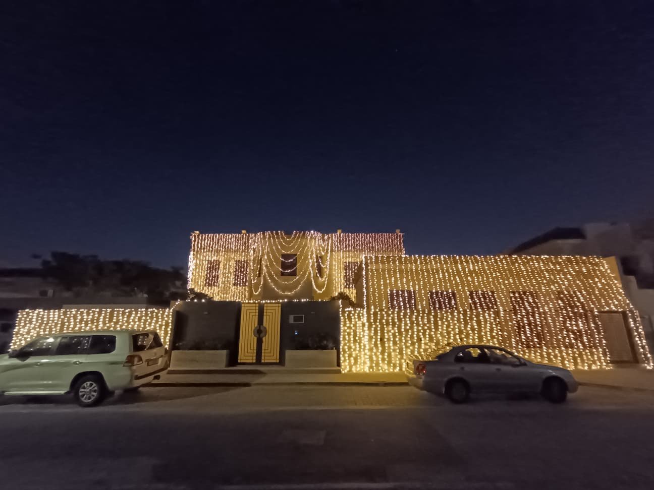 Home house fully decorated with warm golden led curtain lights covering the entire exterior, glowing brightly at night with cars parked in front.